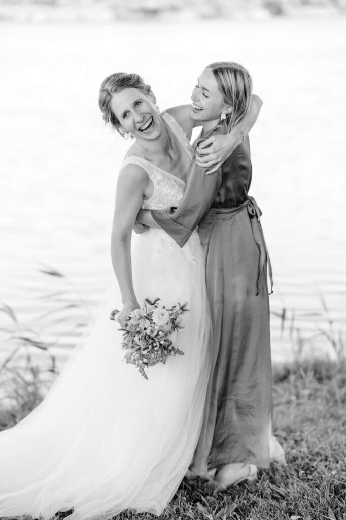 Braut und Trauzeugin umarmen sich lachend am See