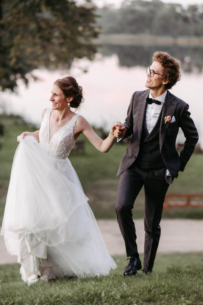 Braut und Bräutigam gehen Hand in Hand vor dem See entlang, festgehalten von der Hochzeitsfotografin Mecklenburg