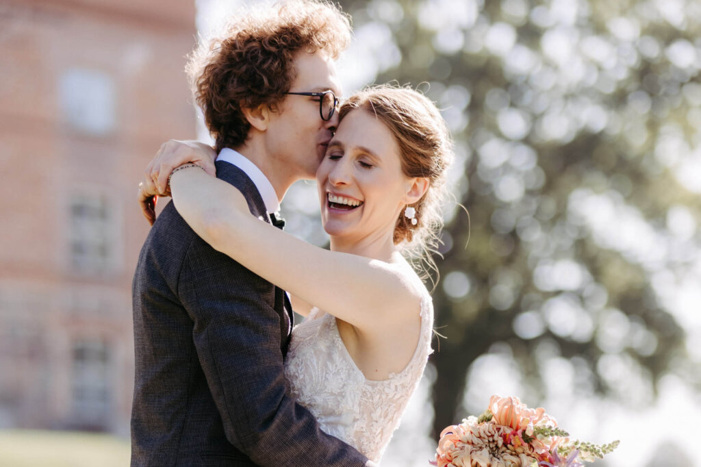 Bräutigam küsst lachende Braut auf die Schläfe vor dem Schloss Ulrichshusen