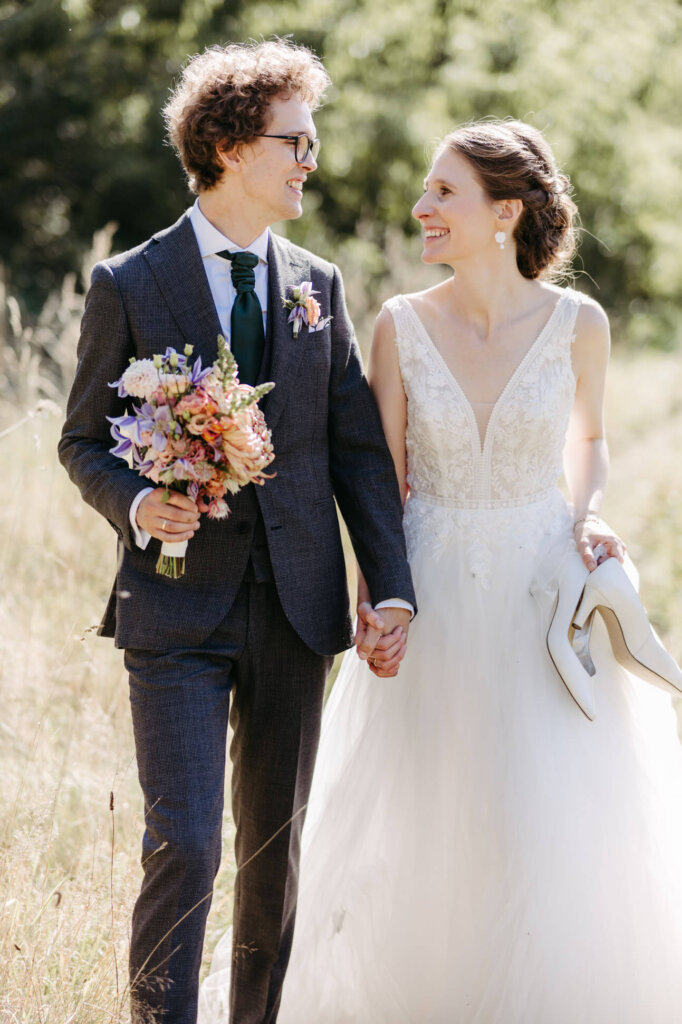 Brautpaar läuft im Sonnenlicht auf einer Wiese entlang, beide lächeln sich an