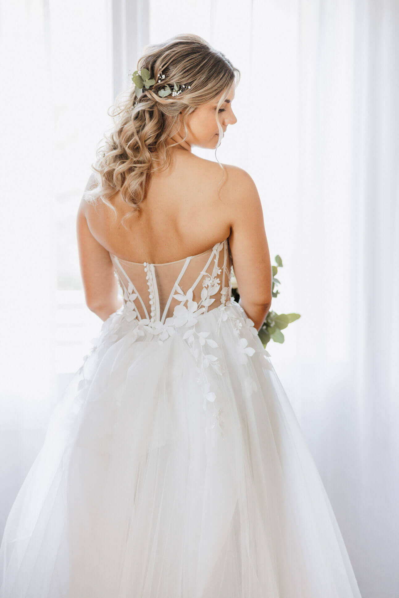 Braut steht vor einem großen hellen Fenster mit dem Rücken zum Fotografen in ihrem Brautkleid, man sieht ihren freien Rücken. High Key Aufnahme