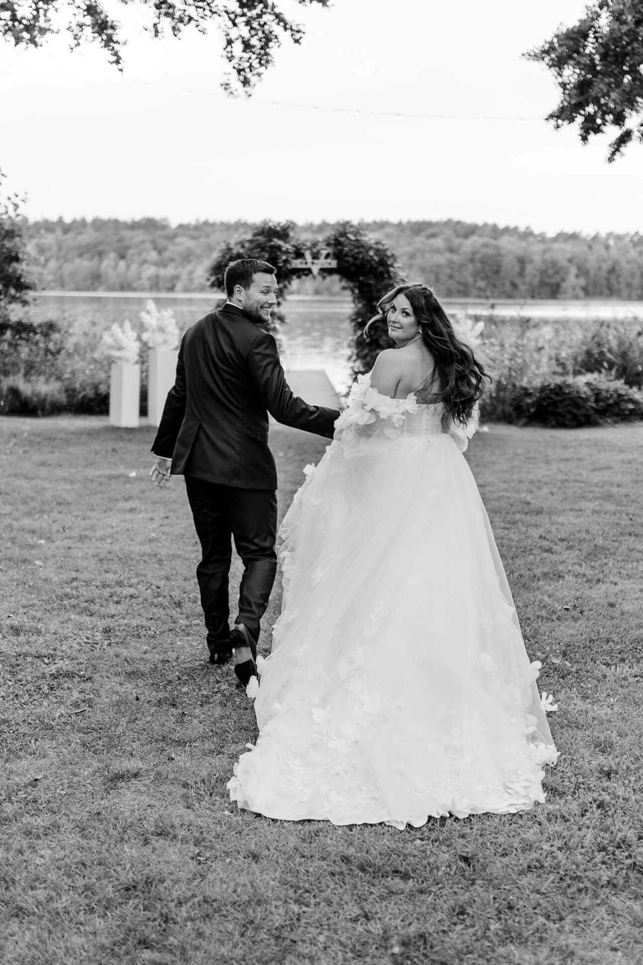Schwarz-weiß-Aufnahme eines Brautpaares, das lachend Hand in Hand Richtung See läuft und dabei in Richtung der Hochzeitsfotografin Mecklenburg und Brandenburg schaut