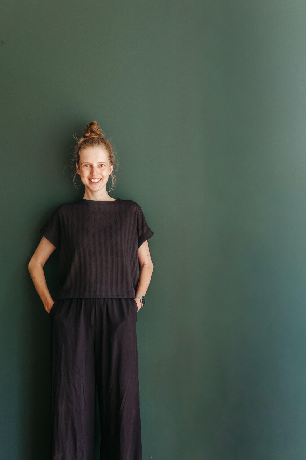Fotografin steht vor grüner Wand, Hände in den Taschen, lächelt in die Kamera